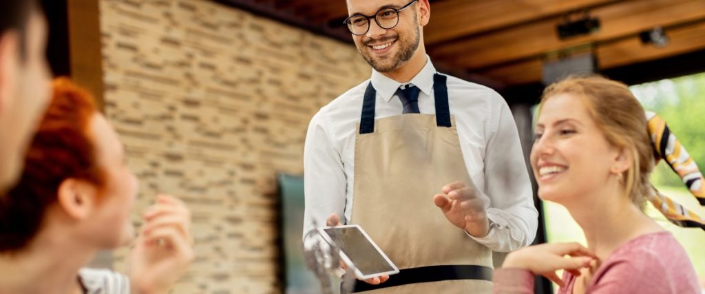 Mesero con delantal toma un pedido en una tablet mientras atiende a clientes en un restaurante.